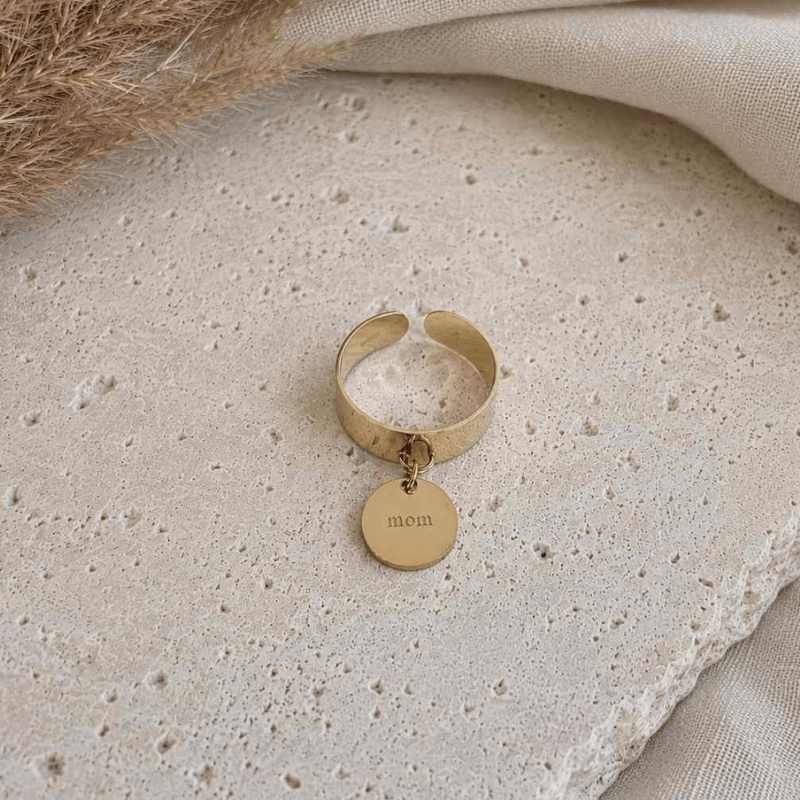 Une bague en acier inoxydable est posée sur une table en travertin, il y a une pampille "mom", bijou de maternité